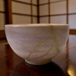 Close-up of a light gray ceramic bowl with a yellow string laid across it, on a dark wooden table (background blurred).