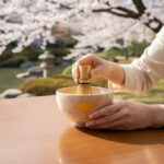 Person whisking matcha in a ceramic crackle-glaze bowl outdoors with cherry blossoms in the background, hands visible.
