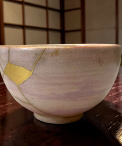 Close-up of a light ceramic bowl on a dark wooden table, with a yellow tape-like mark on its side and a blurred background of shelves.