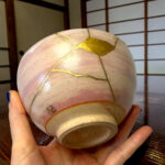 Close-up of a round beige object with gold crack repairs (kintsugi) being held by two hands, wooden floor in the background.