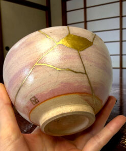 Close-up of a round beige object with gold crack repairs (kintsugi) being held by two hands, wooden floor in the background.