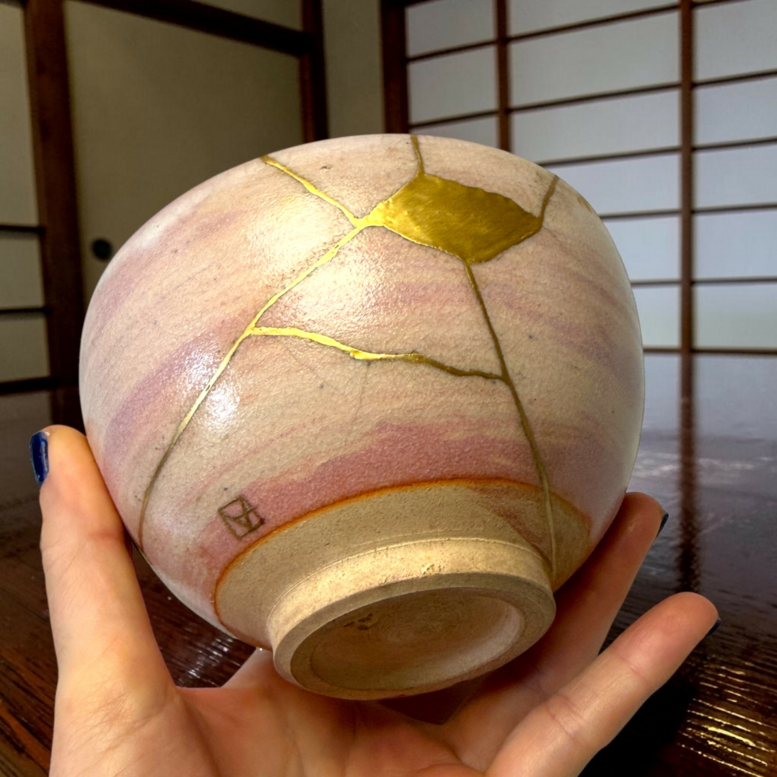 Close-up of a round beige object with gold crack repairs (kintsugi) being held by two hands, wooden floor in the background.