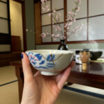 Close-up of a hand holding a blue-and-white ceramic teacup with cherry blossom glaze in a Japanese-style room with a low wooden table and vase of blooms in the background