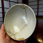 Close-up of a pale ceramic plate with gold repair lines (kintsugi), held by a hand with a wooden surface in the background, showing cracks and glaze.