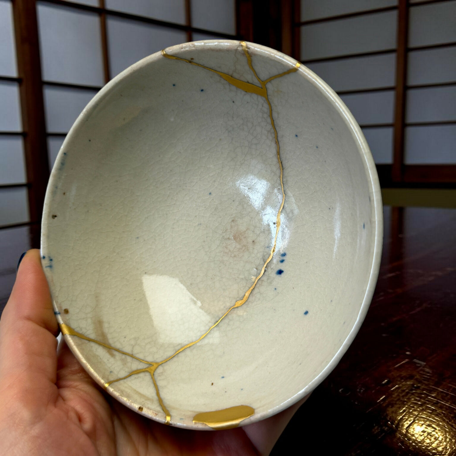 Close-up of a pale ceramic plate with gold repair lines (kintsugi), held by a hand with a wooden surface in the background, showing cracks and glaze.