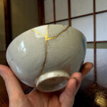 Close-up of a pale ceramic sphere with gold repair seams (kintsugi) being held by hands.