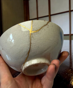 Close-up of a pale ceramic sphere with gold repair seams (kintsugi) being held by hands.