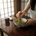 Person whisking bright green matcha with a bamboo chasen in a ceramic bowl on a wooden table.