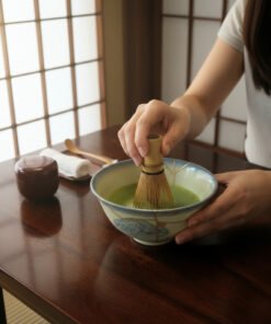 Person whisking bright green matcha with a bamboo chasen in a ceramic bowl on a wooden table.