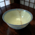 Close-up of a pale blue ceramic bowl with gold repair seams (kintsugi) on a dark wooden table.