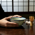 Hands cradle a blue-green ceramic bowl on a dark wooden table, with a bamboo tea whisk nearby and a small towel in the background.