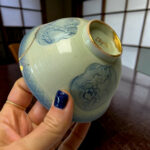 Close-up of a pale blue ceramic vase with floral relief and a gold rim, held by a hand with blue nail polish.