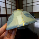 Close-up of a pale blue ceramic lantern with a blue floral pattern, held by a hand with a yellow cord wrapped around it, on a dark wooden surface.