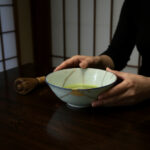 Hands cradle a light blue ceramic bowl with frothy green matcha on a dark wooden table; a bamboo whisk rests nearby.