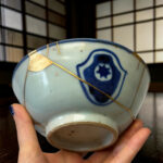Close-up of a ceramic plate with blue patterns and gold kintsugi repair seams, held by hands.