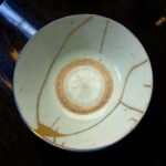 Close-up of a light blue ceramic plate with brown concentric rings and thin gold cords, resting on a dark wooden table.