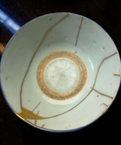 Close-up of a light blue ceramic plate with brown concentric rings and thin gold cords, resting on a dark wooden table.