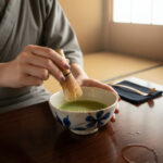 Person whisking matcha in a ceramic bowl with a bamboo chasen, at a wooden table in a sunlit room
