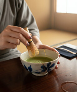 Person whisking matcha in a ceramic bowl with a bamboo chasen, at a wooden table in a sunlit room