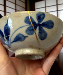 Ceramic bowl with blue glaze patterns showing a gold repaired crack (kintsugi), cradled by small hands against a wooden background.