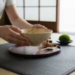 Person’s hands lift a beige ceramic bowl above a red lacquered tray during a Japanese tea ceremony.