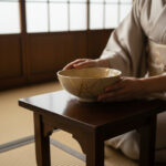 Person in a kimono holds a beige ceramic bowl with gold crackle glaze on a dark wooden table.