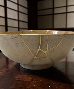 Close-up of a light gray ceramic bowl with beige string repairs resting on a dark wooden table, background shows tiled wall patterns.