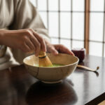 Person uses a bamboo whisk to blend matcha in a ceramic bowl on a wooden table, with soft natural light in the background