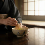 Person wearing a black kimono whisking matcha with a bamboo chasen over a ceramic tea bowl on a wooden table in a traditional setting.
