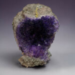 Rough rock with purple amethyst crystals and a gold vein against a gray background, close-up shot