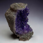 Amethyst crystal cluster attached to a rough gray rock, displaying deep purple, faceted crystals with a pale gradient background