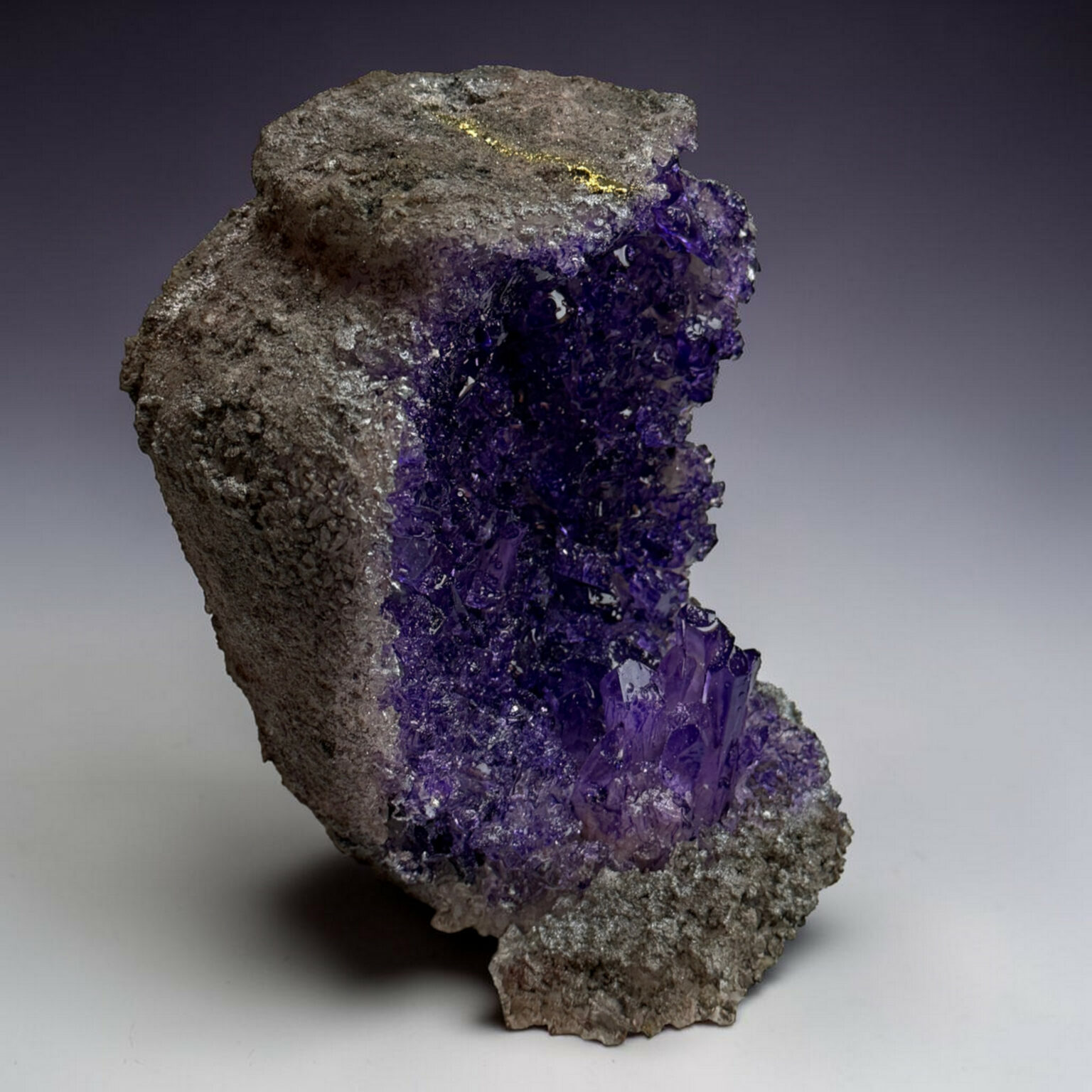 Amethyst crystal cluster attached to a rough gray rock, displaying deep purple, faceted crystals with a pale gradient background