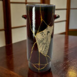 Close-up of a black ceramic vase with gold crackle seams and a painted design, sitting on a wooden table.