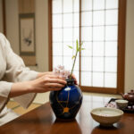 Person in a light robe arranges pink blossoms in a dark blue ceramic vase on a wooden table in a Japanese-style room with shoji screens.