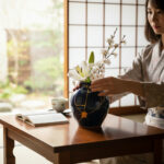Woman in a kimono arranges white lilies and cherry blossoms in a blue vase on a wooden table by a shoji screen in a sunlit room.
