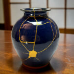 Blue ceramic vase with gold kintsugi cracks on a wooden table surface.