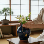 Hands arranging white lilies in a glossy dark blue vase with gold crackle pattern on a wooden table near a bonsai under a shoji-style window inside.
