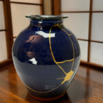 Close-up of a dark blue ceramic vase with gold kintsugi cracks on a wooden table surface.