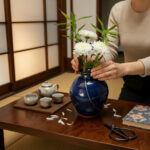 Person arranging white chrysanthemums in a dark blue vase on a wooden table, with a tea set and scissors nearby in a cozy room.