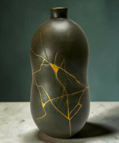 Dark ceramic vase with gold repair seams (kintsugi) on a gray floor against a green wall; vertical, close-up shot.