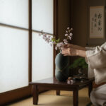 Person in a light robe arranges pink blossoms in a dark green vase on a low wooden table in a Japanese-style room with a shoji screen in the background.