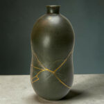 Close-up of a dark ceramic vase with thin gold wraps running diagonally down its surface, set against a gray backdrop on a concrete floor.