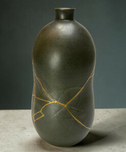 Close-up of a dark ceramic vase with thin gold wraps running diagonally down its surface, set against a gray backdrop on a concrete floor.
