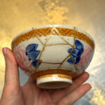 Close-up of a hand holding a ceramic bowl with gold-kintsugi cracks and blue floral glaze, against a warm background.