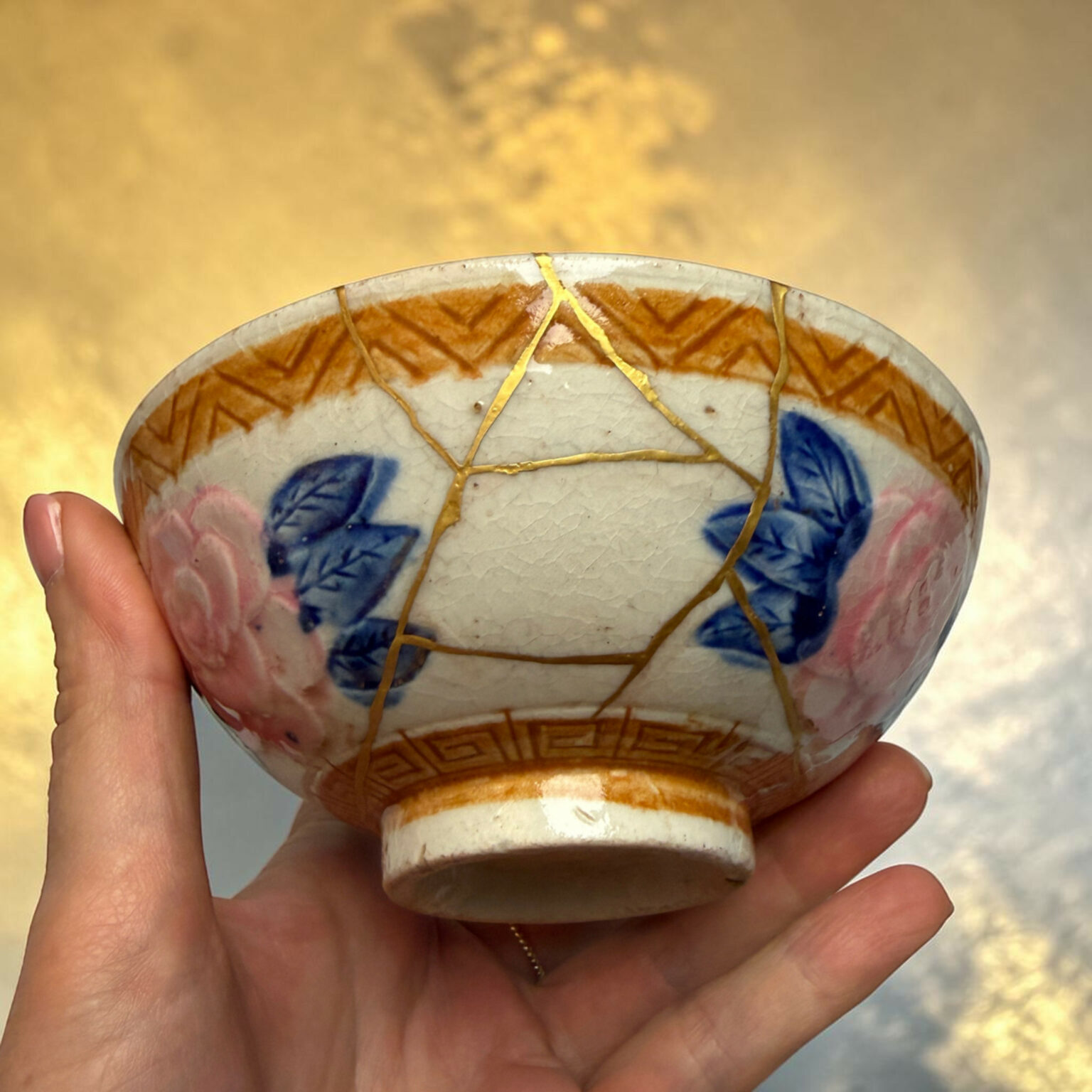 Close-up of a hand holding a ceramic bowl with gold-kintsugi cracks and blue floral glaze, against a warm background.
