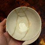 Close-up of a ceramic bowl with gold kintsugi cracks, held by a hand against a dark wooden background.