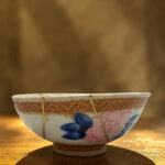 Decorative ceramic hanging bowl with blue and pink floral pattern, suspended by strings against a warm blurred background.