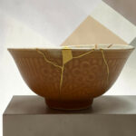 Close-up of a brown ceramic pot with yellow straps on a light pedestal, against a pale geometric background with a triangle shape.
