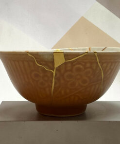 Close-up of a brown ceramic pot with yellow straps on a light pedestal, against a pale geometric background with a triangle shape.