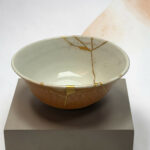 Ceramic bowl repaired with gold kintsugi on a gray display plinth against a light background.